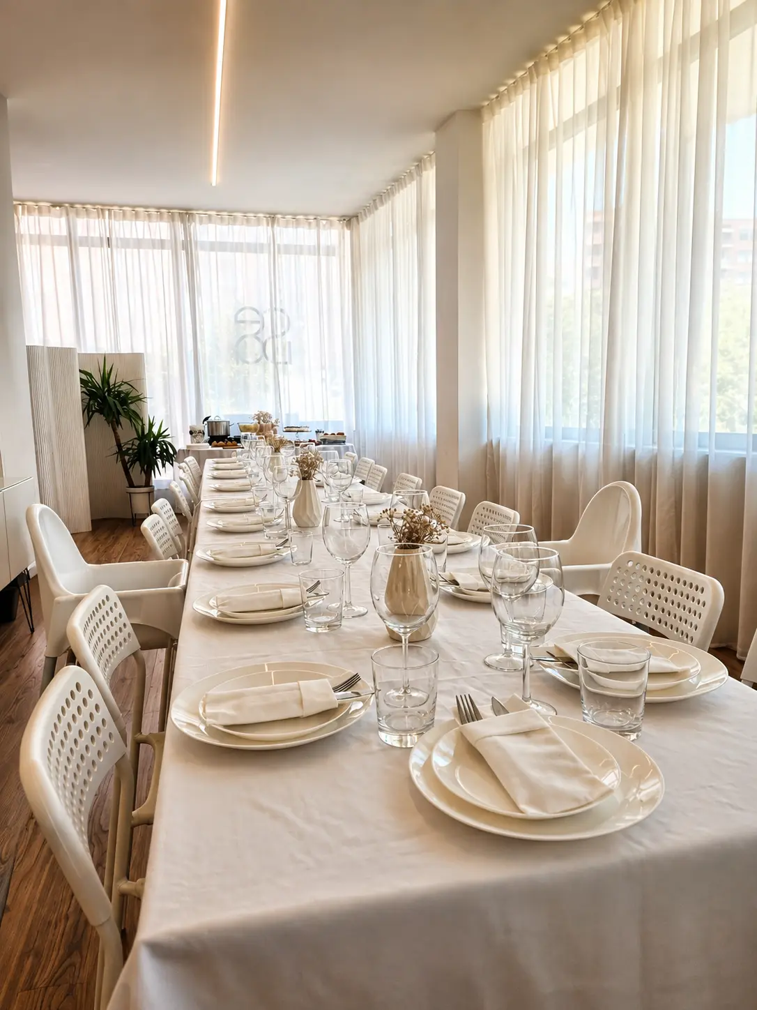 Mesa comprida posta para jantar, com loiça branca, copos de vinho e luz natural