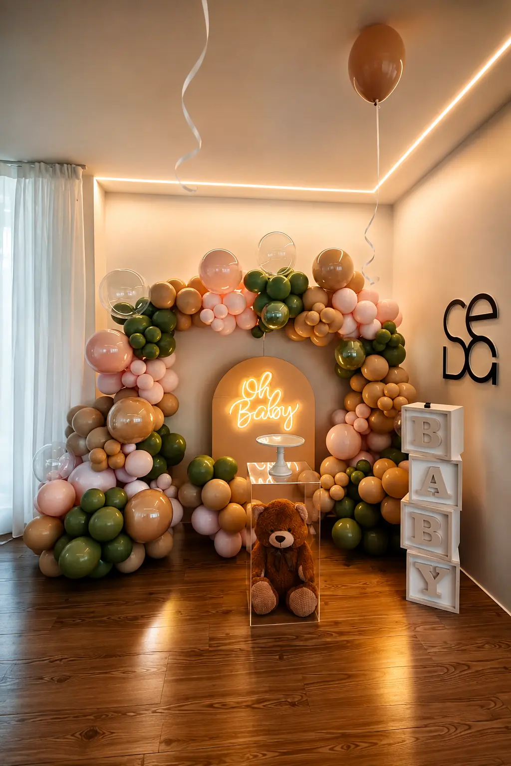 Cenário de baby shower com arco de balões em tons rosa, verde e caramelo e letreiro luminoso Oh Baby